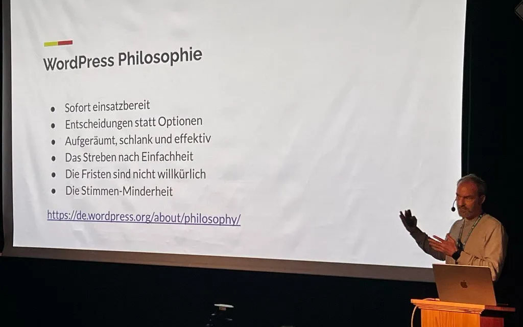 Torsten Landsiedel auf der Bühne des WordCamp Leipzig. Er hat ein Headset auf und zeigt auf eine Folie hinter ihm.